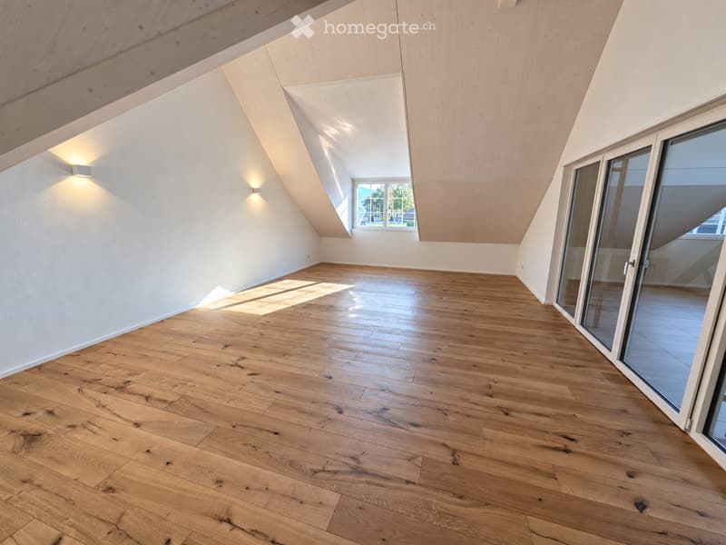 Empty room with wooden flooring, white walls, and large windows allowing natural light. The ceiling has a slanted design with built-in lighting.