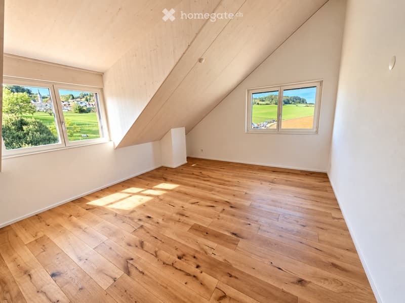 Bright, empty attic room with wooden flooring, white walls, and large windows offering a scenic view of green fields and a village.