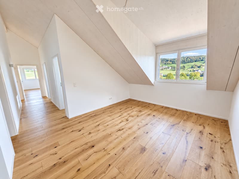 Bright, empty room with wooden flooring and white walls, featuring large windows with scenic views and a door leading to another space.