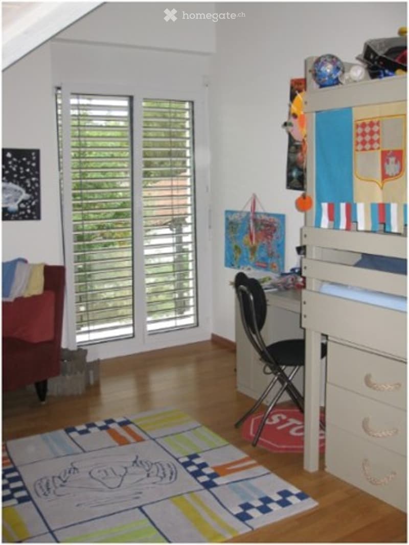A cozy, well-lit room with a wooden floor, large windows with blinds, a red sofa, a colorful rug, a desk with a chair, and a storage unit with drawers.