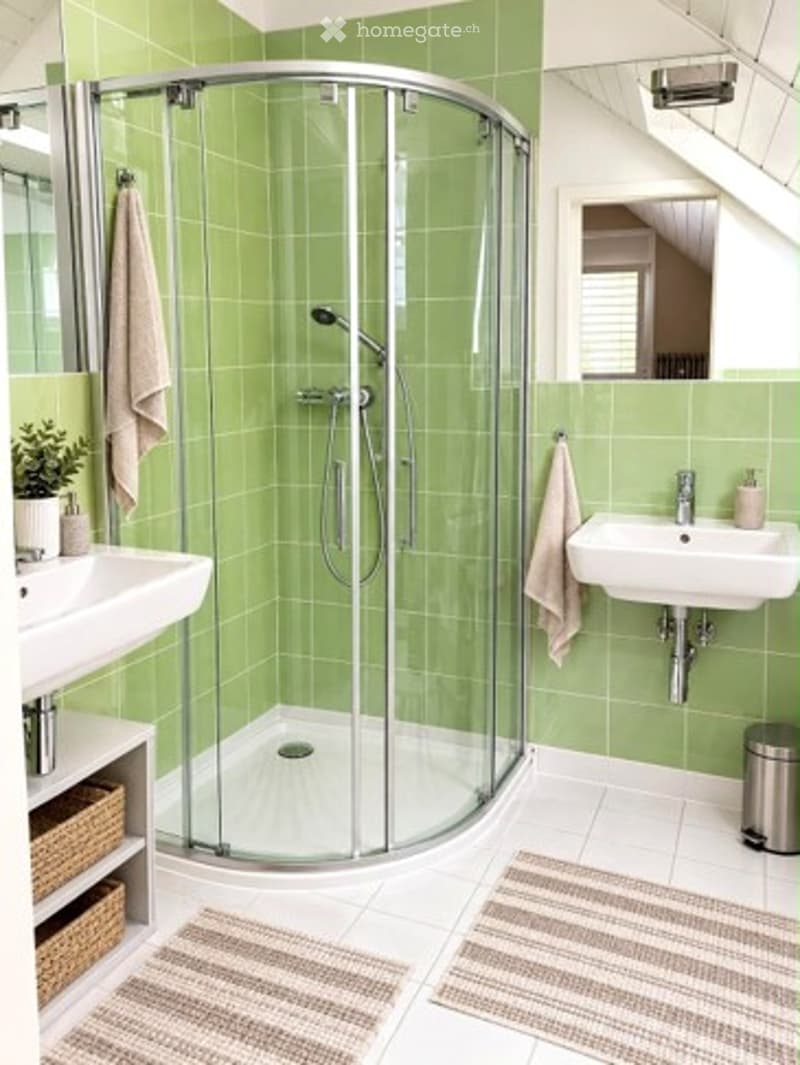 Modern bathroom with green tiled walls, white floor, glass-enclosed shower, two sinks, towels, small plant, and wicker baskets.