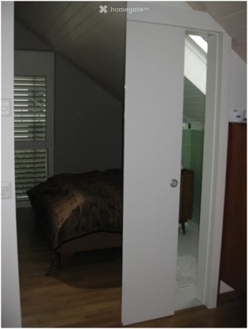 A cozy bedroom with wooden flooring, a bed with a patterned comforter, a nightstand, and a partially open door leading to a bathroom with green tiles.