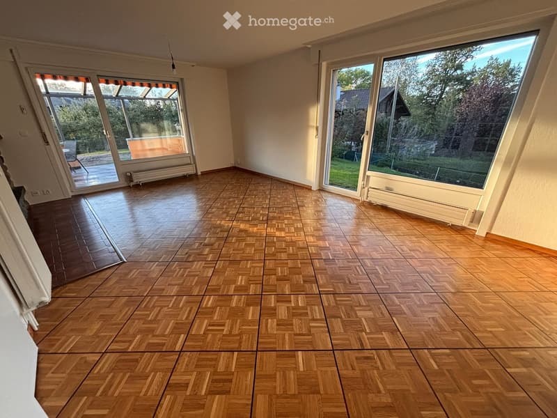 Empty room with wooden herringbone flooring, large windows, and sliding glass doors leading to an outdoor area.