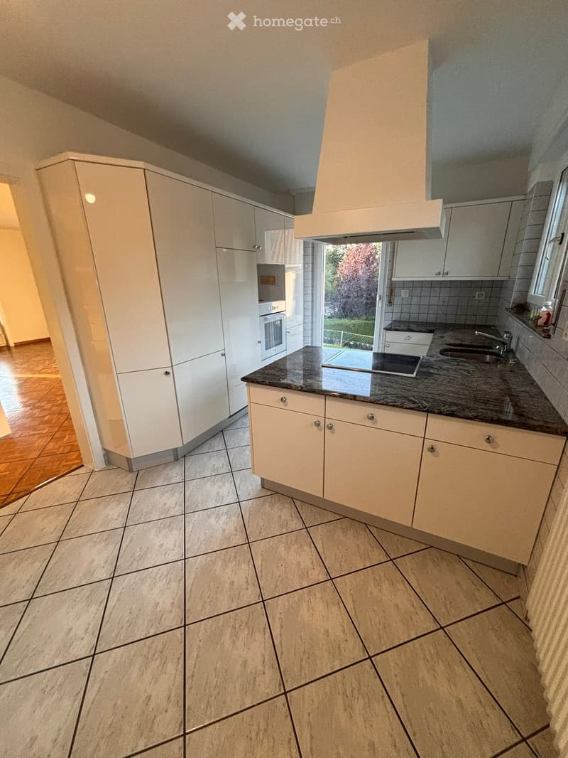 Modern kitchen with white cabinets, granite countertop, stainless steel sink, and built-in oven. Features tiled floor and large window with view of greenery.