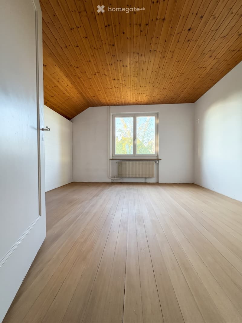 Empty room with light wooden flooring, white walls, and a slanted wooden ceiling. Features a window with a radiator below and a door on the left side.