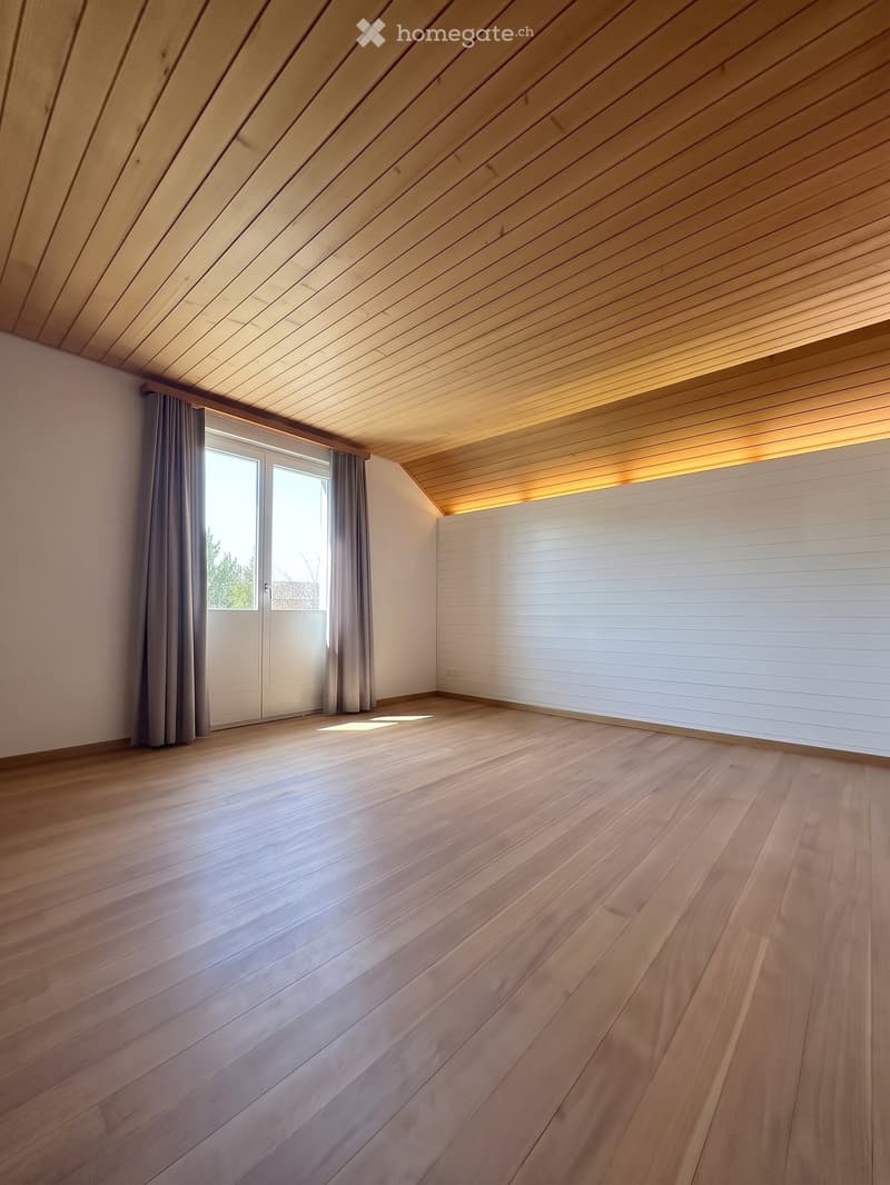 Empty room with wooden flooring, white walls, and large windows with curtains. The ceiling has a wooden slatted design.