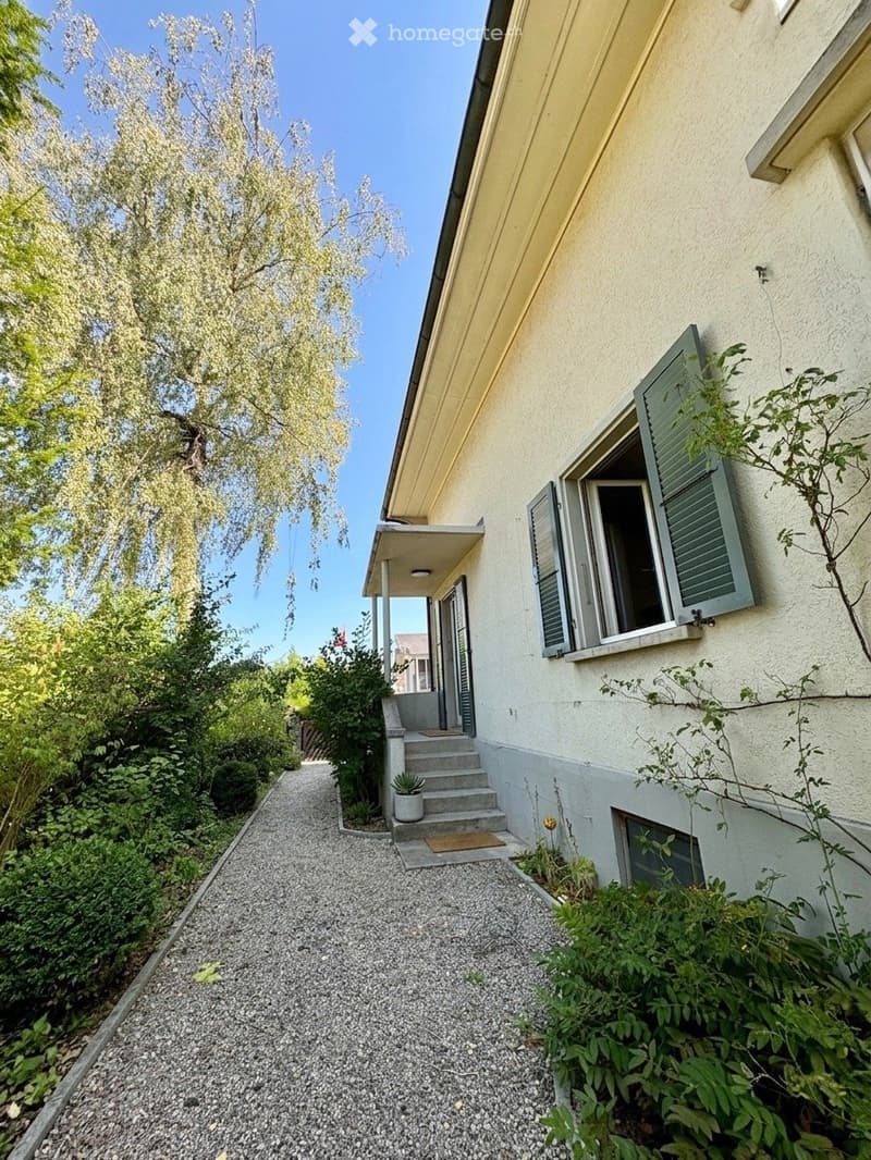 Two-story house with light yellow facade, green shutters, and a small porch. The entrance features a gravel pathway lined with greenery and a potted plant near the steps.