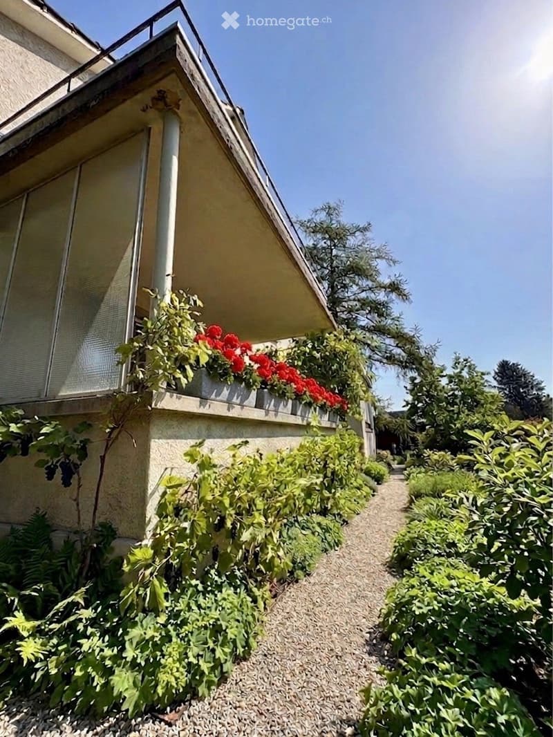 Balcony with red flowers and greenery, surrounded by a garden with gravel path and various plants.