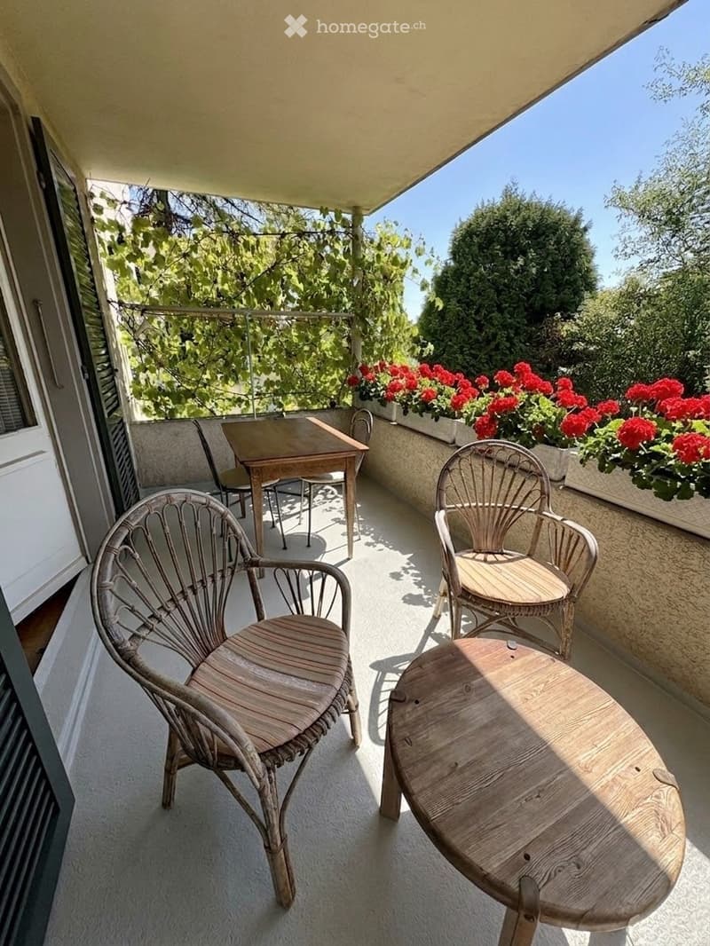 Balcony with wooden table and wicker chairs, surrounded by greenery and red flowers in planters.
