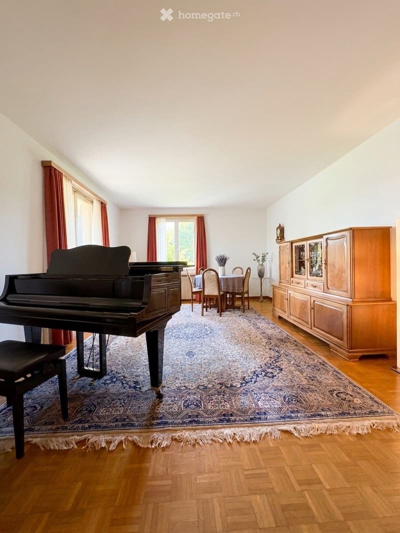 Spacious room with wooden flooring, large area rug, grand piano, dining area with table and chairs, wooden cabinet, and large windows with curtains.