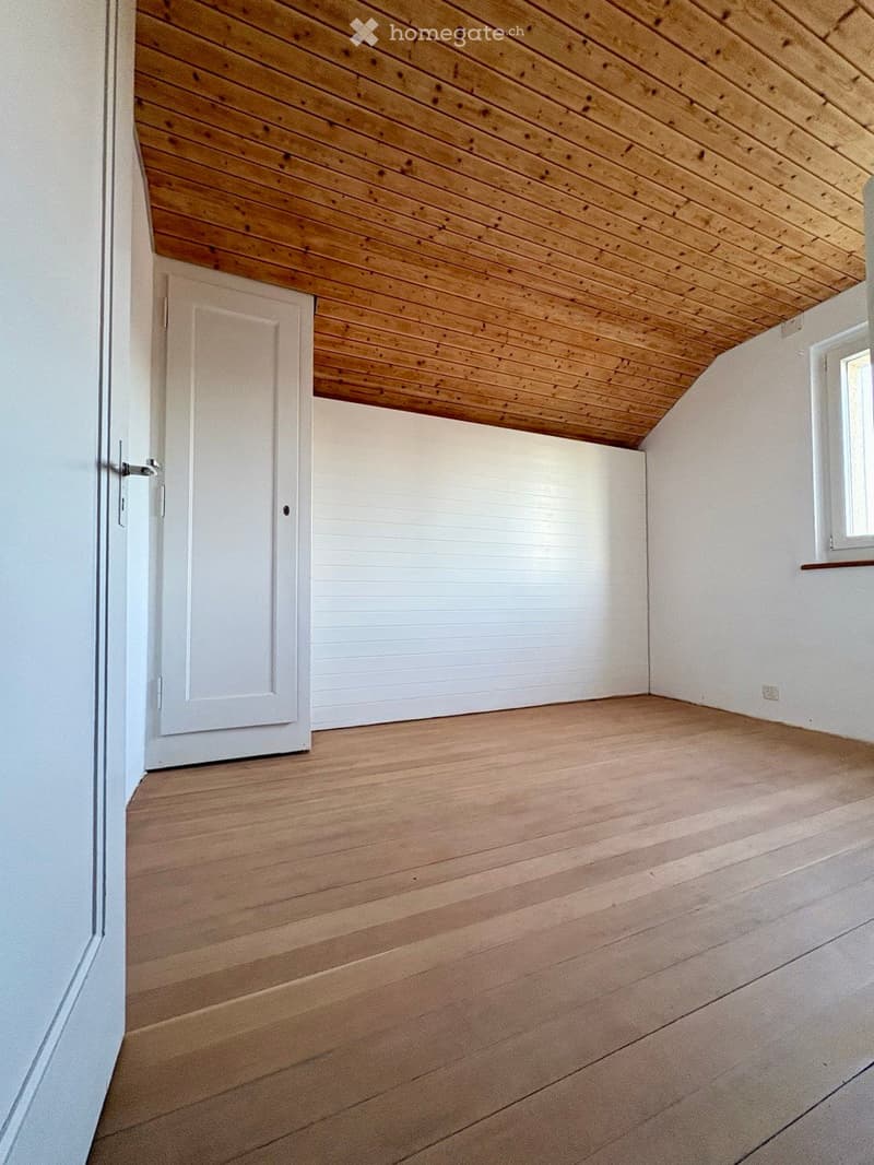 Empty room with wooden flooring, white walls, and a wooden slanted ceiling. Features a white door and a small window with a shelf.