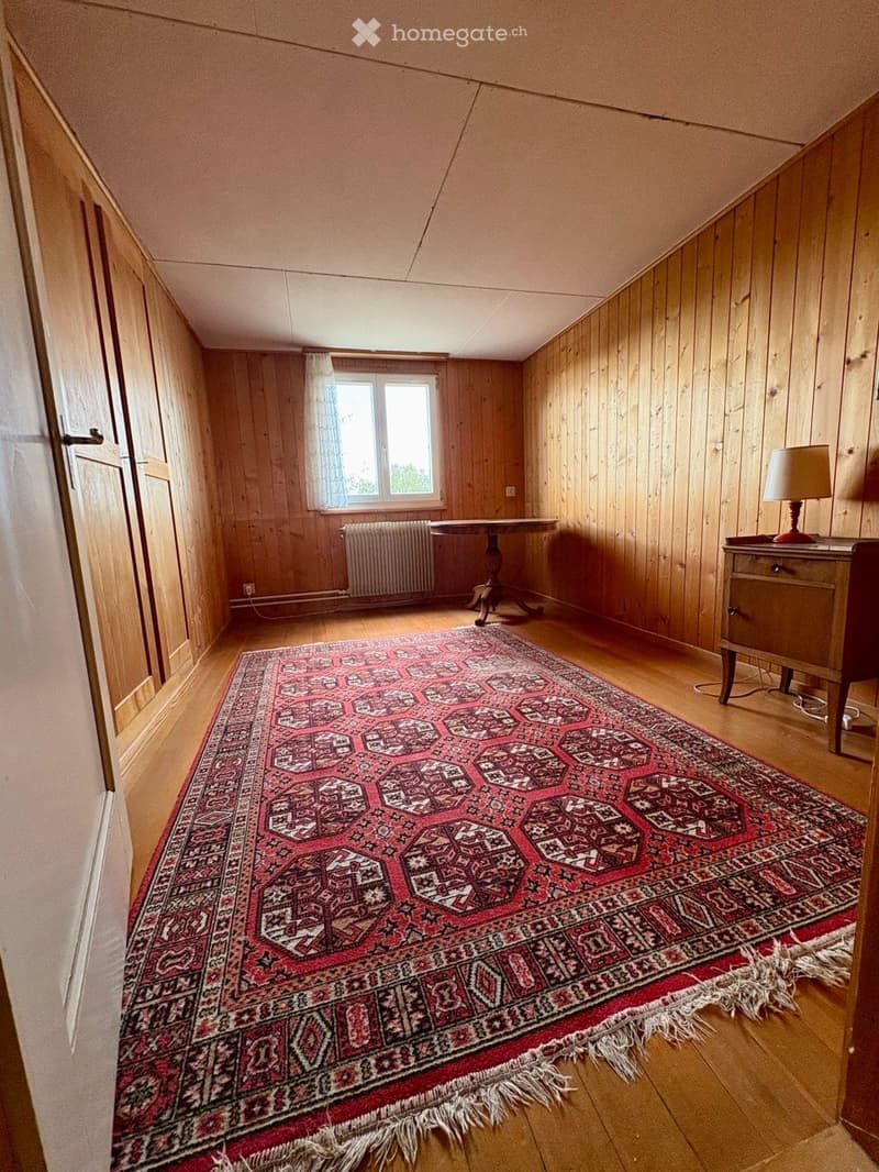 A small, wooden-paneled room with a slanted ceiling, featuring a large patterned carpet, a small table, a lamp, and a radiator under a window.