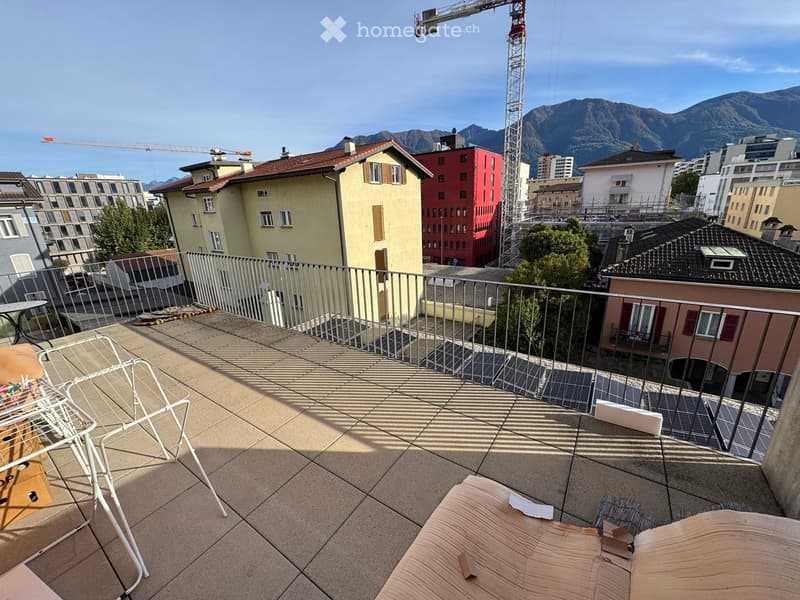 Balcony with a view of city buildings and mountains, featuring a tiled floor, metal railing, and a few pieces of furniture including a chair and a table.
