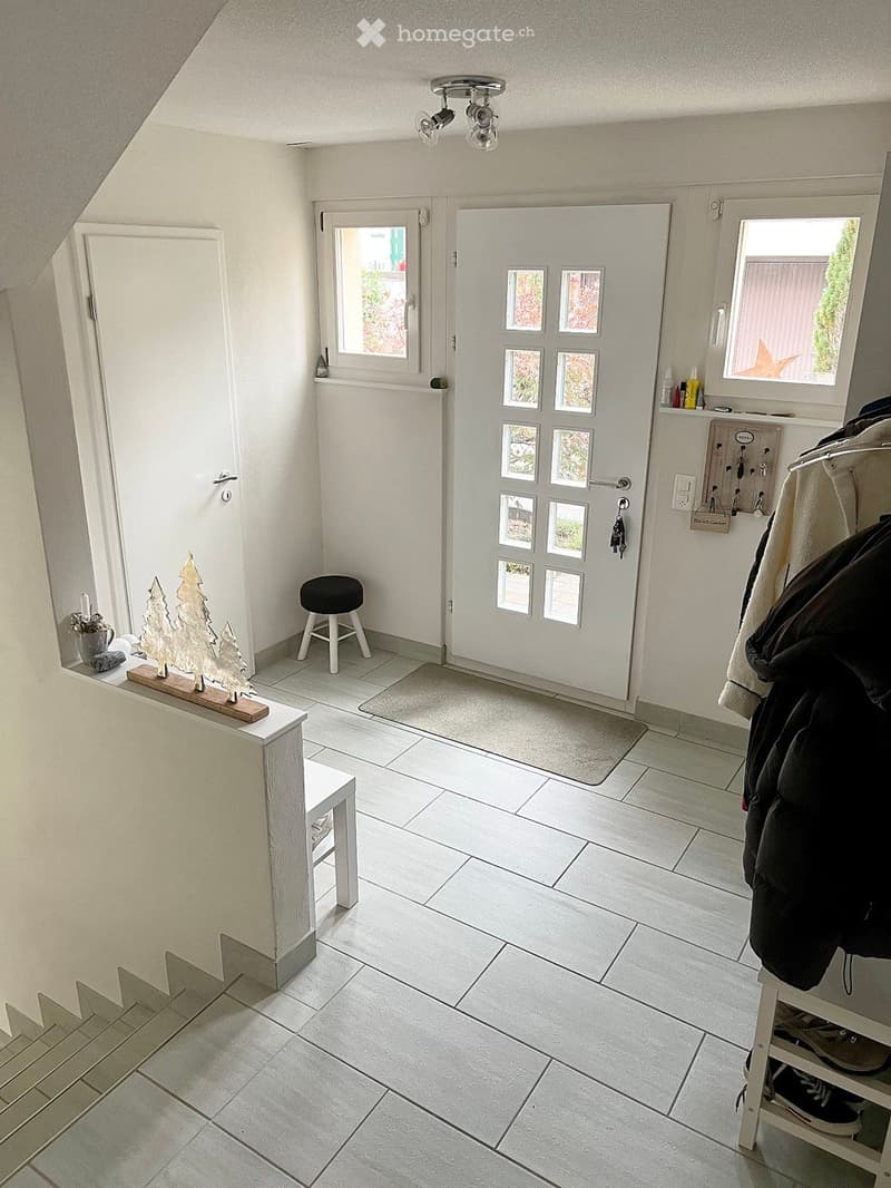Bright hallway with tiled floor, white walls, and a coat rack holding jackets. A small stool sits near the entrance, and a decorative wooden tree sits on a small table near the window.