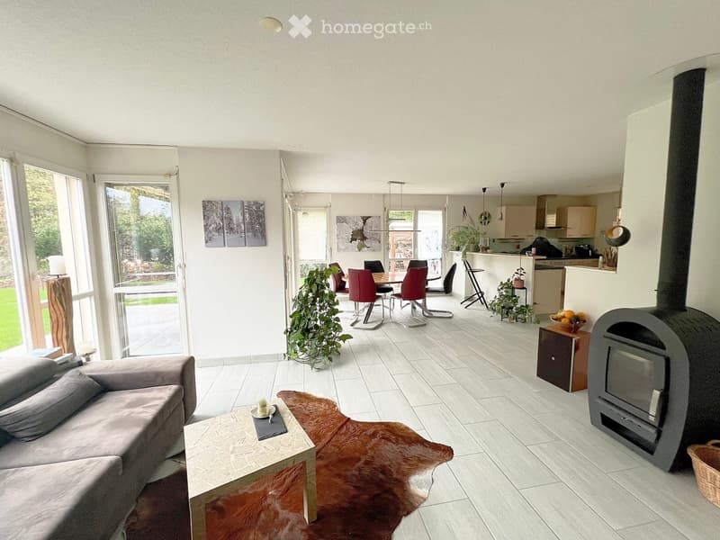 Bright, open-plan living and dining area with white walls, large windows, a grey sofa, coffee table, dining table with red chairs, kitchen area, fireplace, and decorative plants.