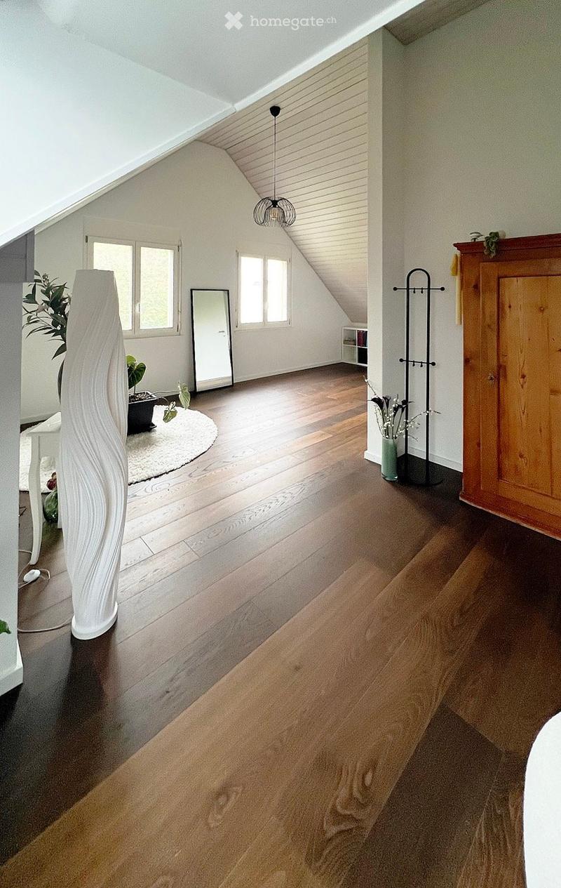 Bright, airy attic room with wooden flooring, large windows, a tall white vase, a round white rug, a mirror, a coat rack, and a small cabinet.