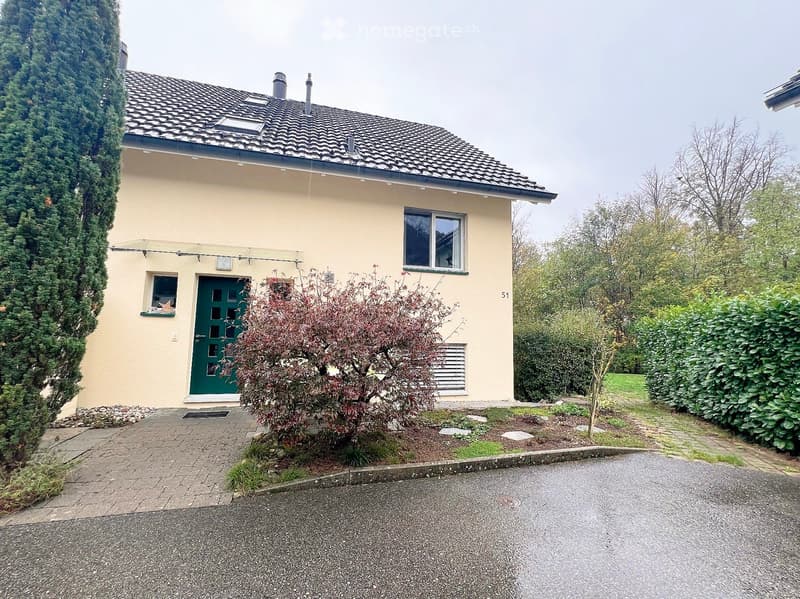 Single-story house with beige exterior, green front door, and small garden area with shrubs and trees.