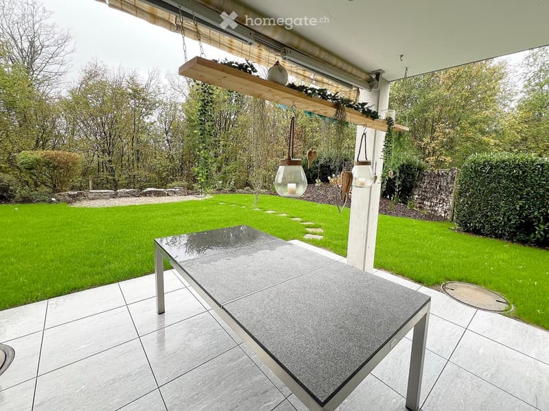 Outdoor patio with a modern grey table, hanging lanterns, and a well-maintained lawn with trees and shrubs.