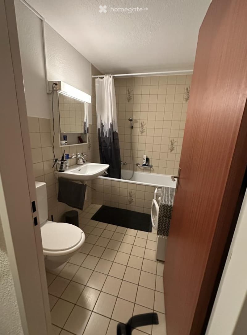 A small bathroom with a toilet, sink, bathtub, and washing machine. The space features tiled walls and floor, a mirror above the sink, and a shower curtain in the bathtub area.