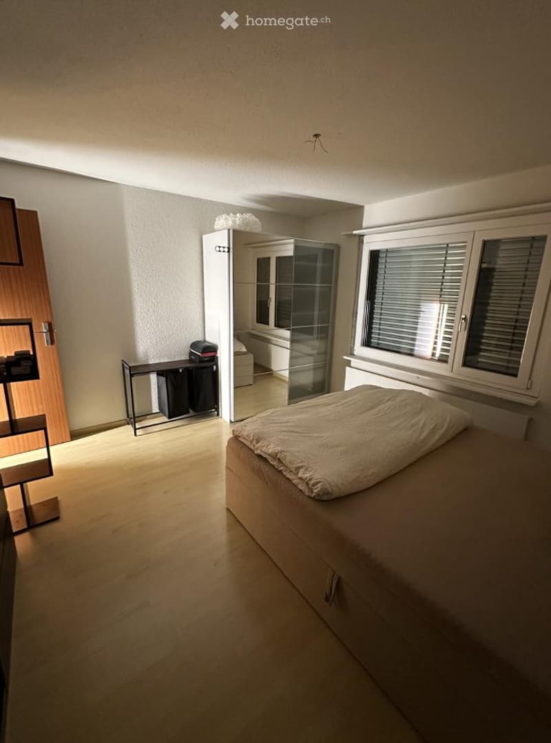 A modern bedroom with a bed, wardrobe, mirror, and a small table with a chair. The room features light wooden flooring and white walls.