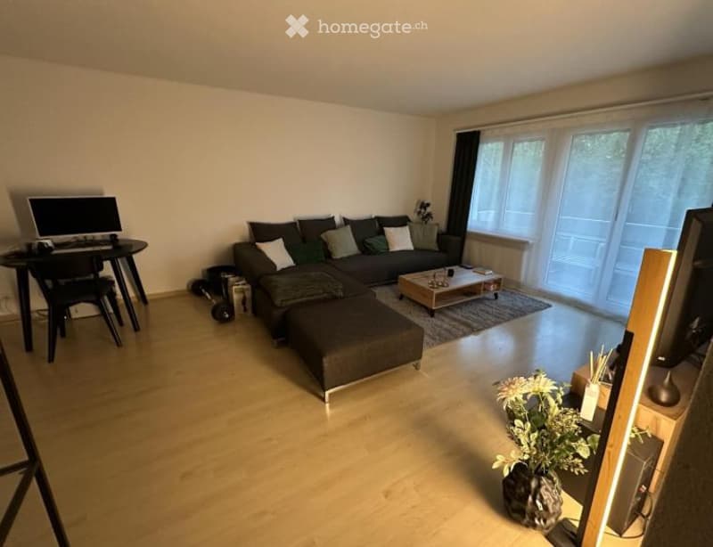 Modern living room with a grey sectional sofa, coffee table, small desk with laptop, and large windows with blinds.