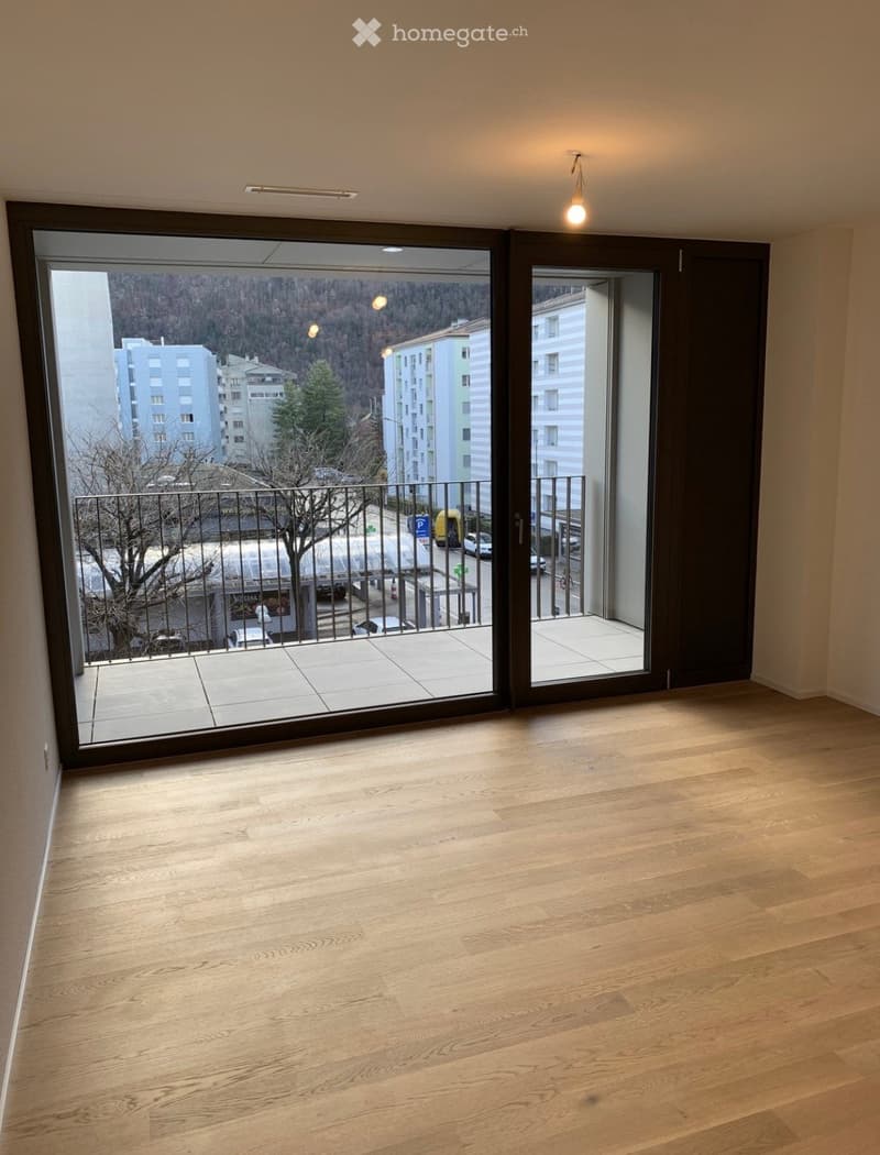 Empty room with wooden flooring and large sliding glass doors leading to a balcony with a view of the city and surrounding buildings.