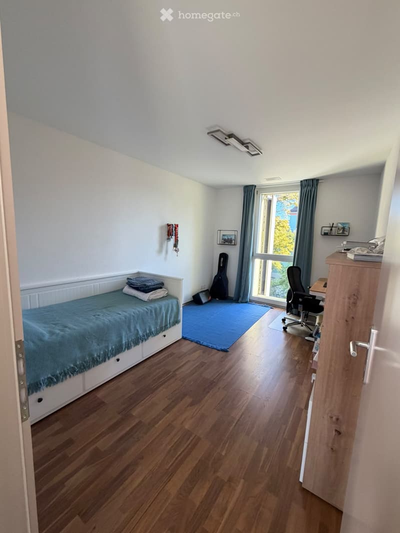 A small, well-lit bedroom with a single bed, a chair, a guitar, a small table, and a window with curtains.
