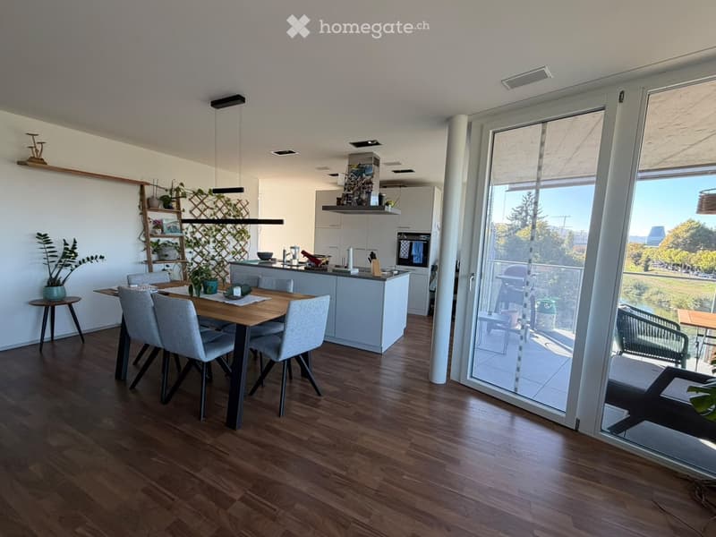 Open-plan kitchen and dining area with wooden flooring, modern white cabinets, a central island, and a dining table with four chairs. Large windows offer a view of the outdoor area.