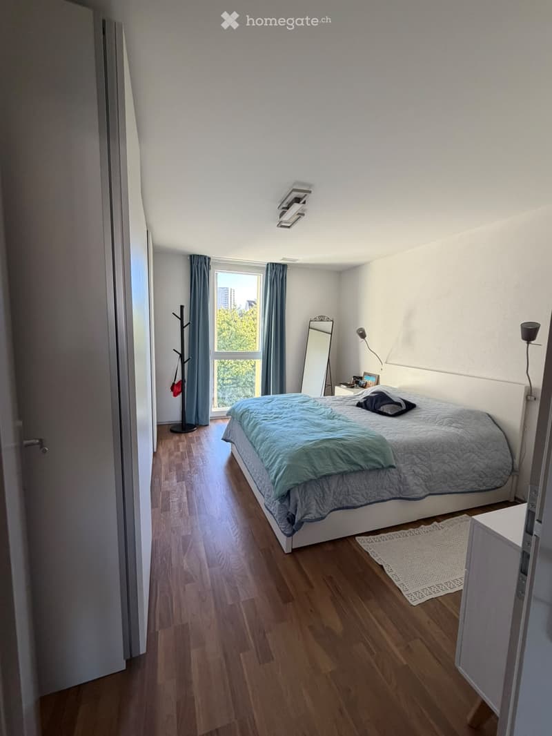 Cozy bedroom with wooden flooring, a large bed with blue bedding, a nightstand with a lamp, a mirror, a coat rack, and a small rug near the bed.