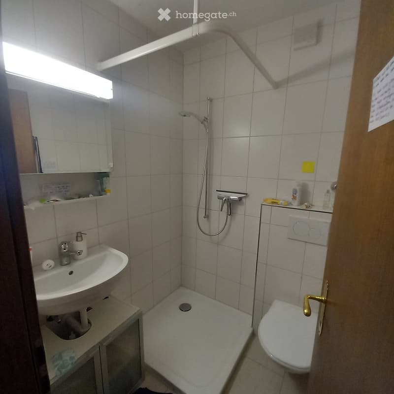 Small bathroom with a sink, toilet, shower, and mirror. The walls are tiled, and there is a small cabinet next to the sink.