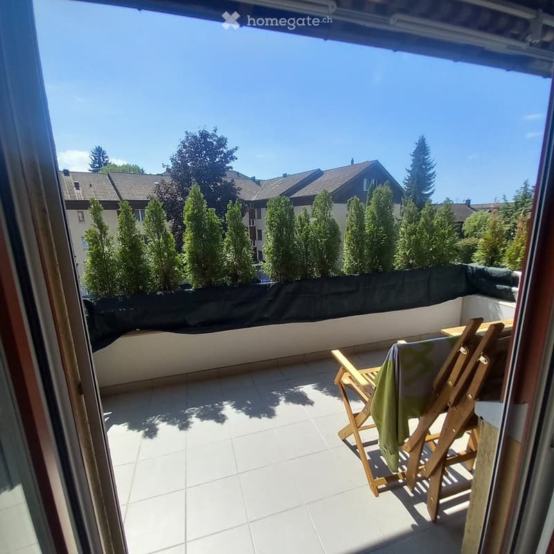 Balcony with tiled floor, wooden chair, small table, and a view of surrounding houses and trees.