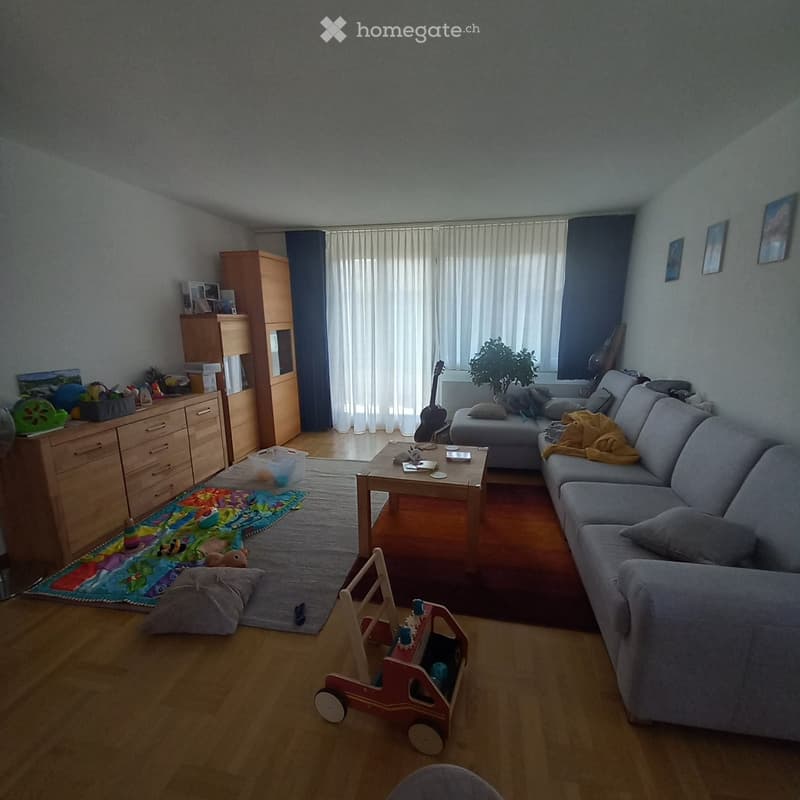 Living room with a grey sofa, coffee table, guitar, plant, cabinet, and toys including a toy car and a colorful play mat.