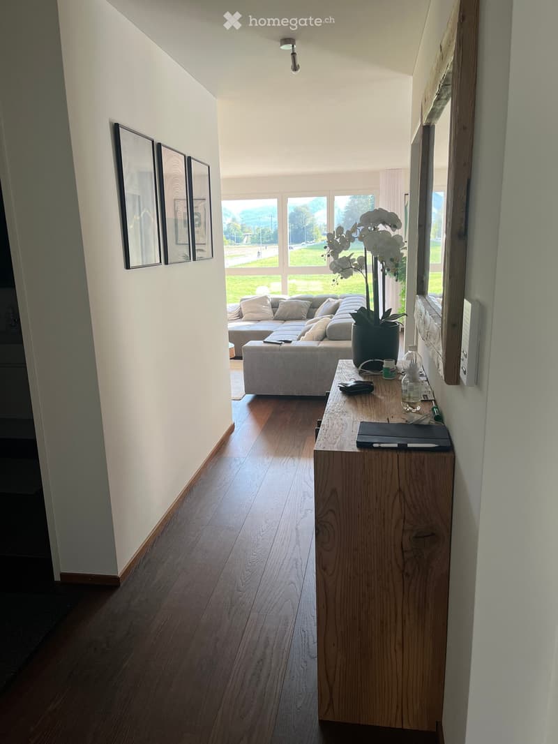 Modern hallway leading to a living area with a sectional sofa, ottoman, wooden table with vase, and framed pictures on the wall.