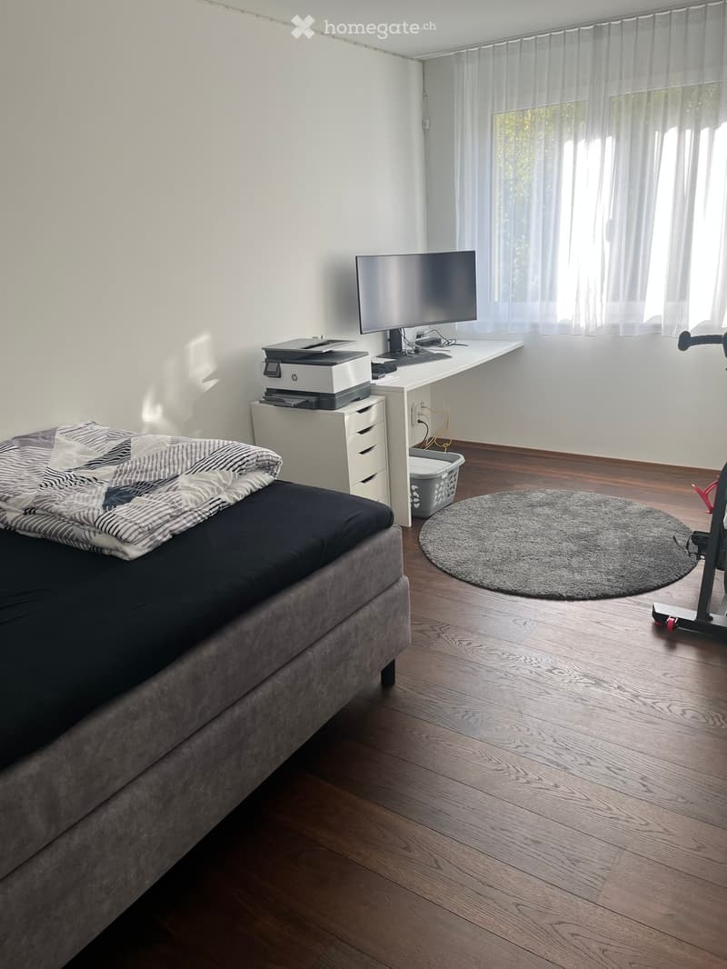 A modern bedroom with a large bed, a desk with a monitor, printer, and keyboard, a round rug, and a scooter in the corner.
