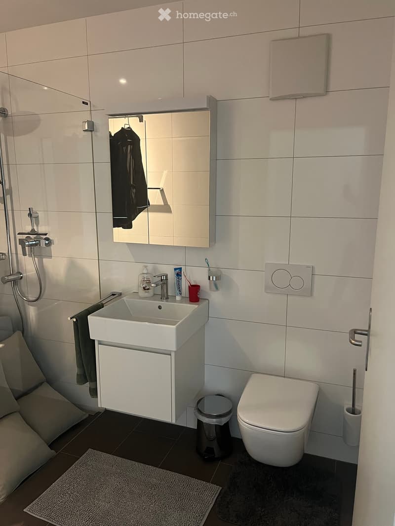 Modern bathroom with white tiles, a sink with a mirror cabinet, a toilet, a towel, and personal care items on the sink counter.
