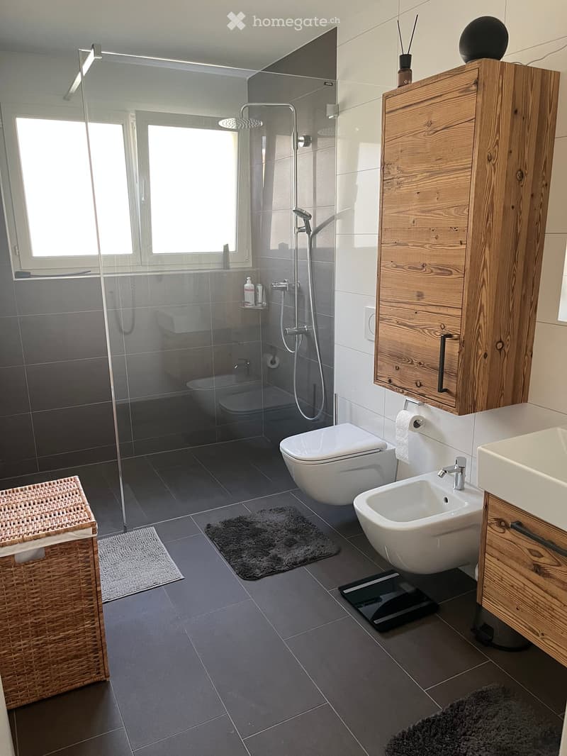 Modern bathroom with dark grey tiles, a wooden cabinet, a toilet, a sink, a shower, and a wicker basket on the floor.