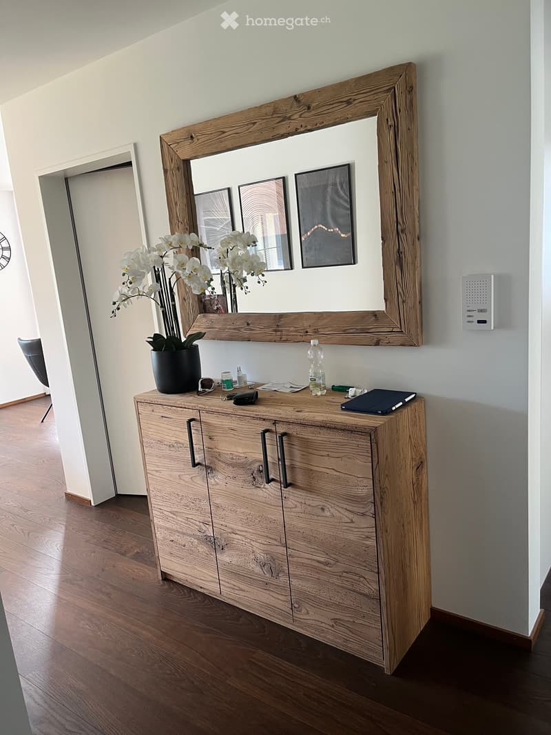 A modern hallway with a wooden cabinet, a large mirror with a wooden frame, a vase with white flowers, and a bottle on the cabinet.