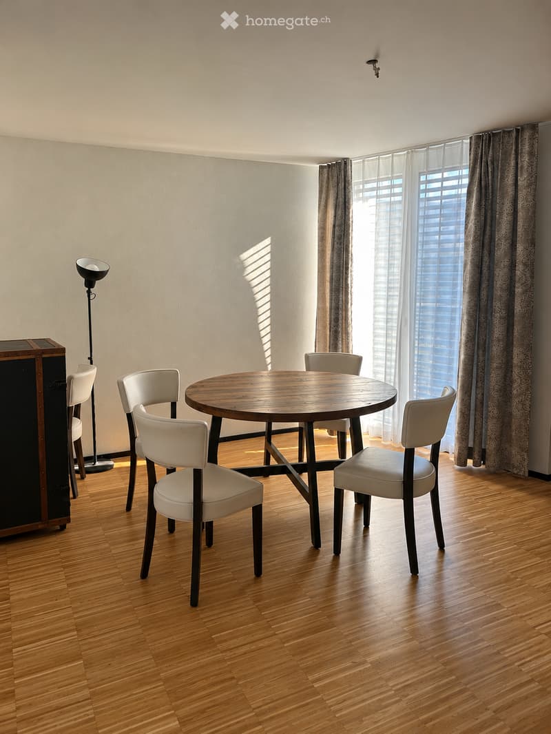 Bright dining area with wooden floor, round wooden table surrounded by six white chairs, floor lamp, and large window with curtains.