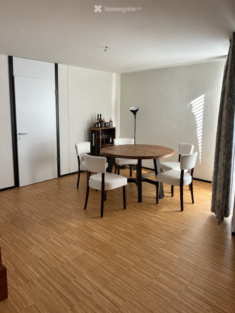 A modern dining area with a round wooden table surrounded by white chairs, a small wooden shelf with bottles, and a floor lamp near a large window with curtains.