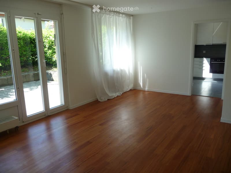 Empty room with wooden flooring, large sliding glass door leading to a patio, and a view of greenery outside. The room has white walls and a radiator on the left side.