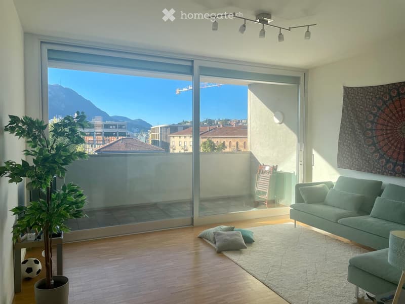 Modern living room with large glass sliding doors leading to a balcony, offering a scenic mountain view. Features a green sofa, throw pillows, a potted plant, and a decorative wall hanging.