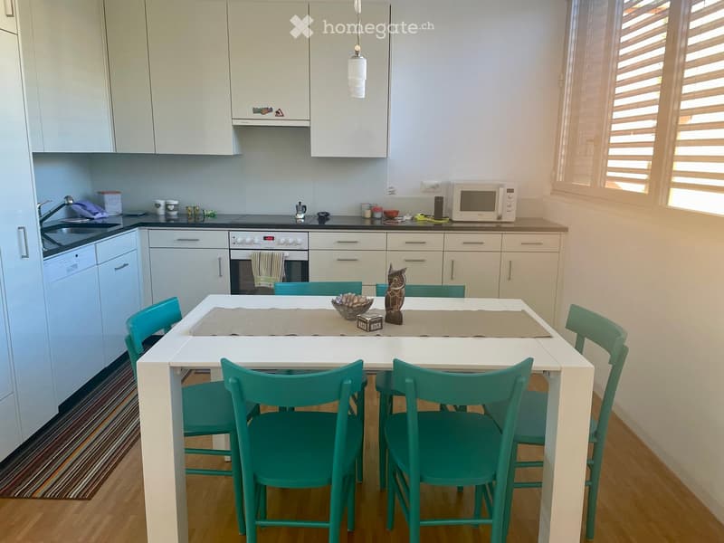 Modern kitchen with white cabinets, a central dining table with green chairs, microwave, oven, and dishwasher. The table is set with a decorative bowl and a small statue.