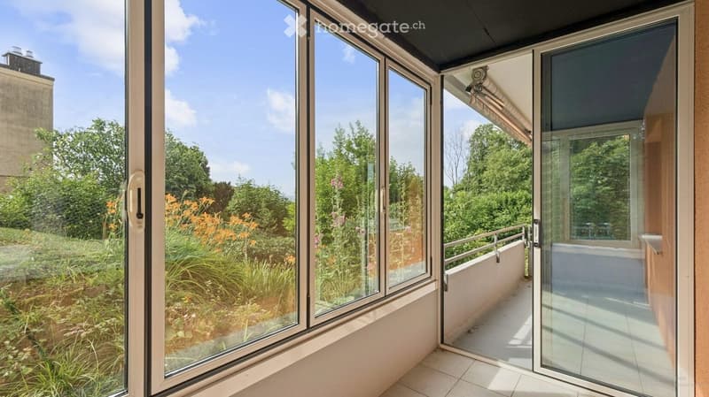 Balcony with large glass doors opening to a lush garden, featuring a metal railing and tiled floor.