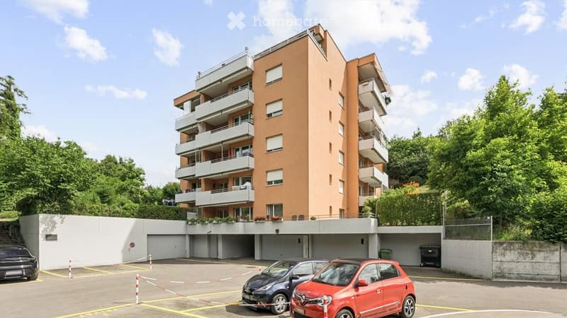 Modern multi-story apartment building with balconies, surrounded by greenery, with a parking area and several cars.