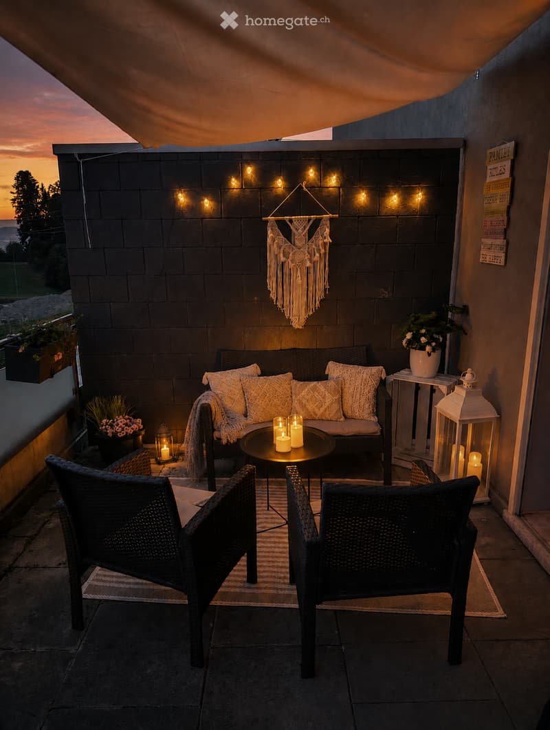 Cozy balcony with a sofa, small round table, two wicker chairs, candles, a lantern, and a decorative hanging ornament.