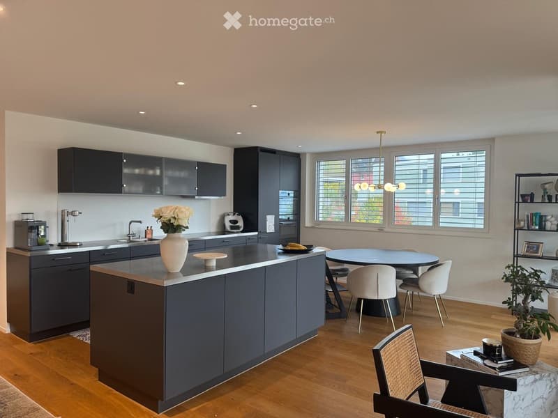 Modern kitchen with dark grey cabinets, a central island, and a dining area with a round table and chairs. Features include a coffee maker, vase with flowers, and a bookshelf with decorative items.