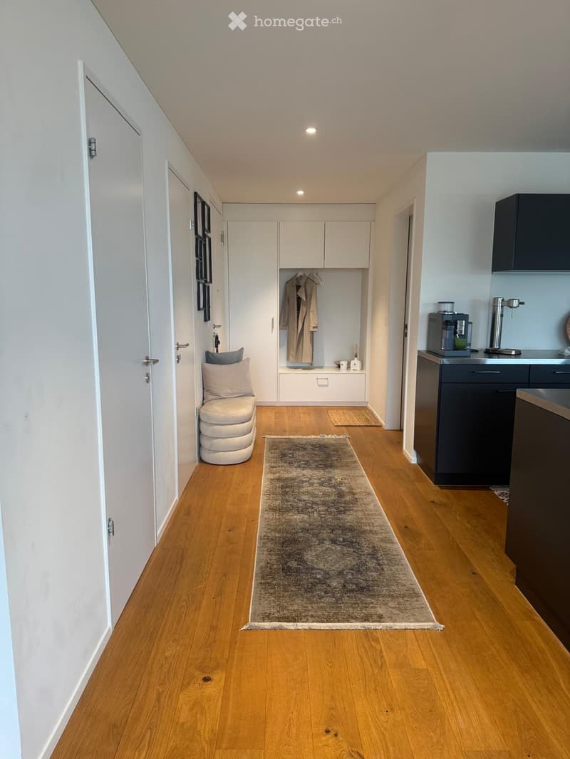 Modern hallway with light wooden flooring, white walls, and built-in storage. Features a small seating area with a stool, a coffee machine on a counter, and a coat hanging on a rack.