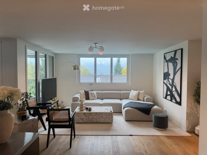 Modern living room with light-colored sofa, coffee table, chairs, and decorative items. Large windows provide natural light, complemented by a floor lamp and wall-mounted lighting fixture.