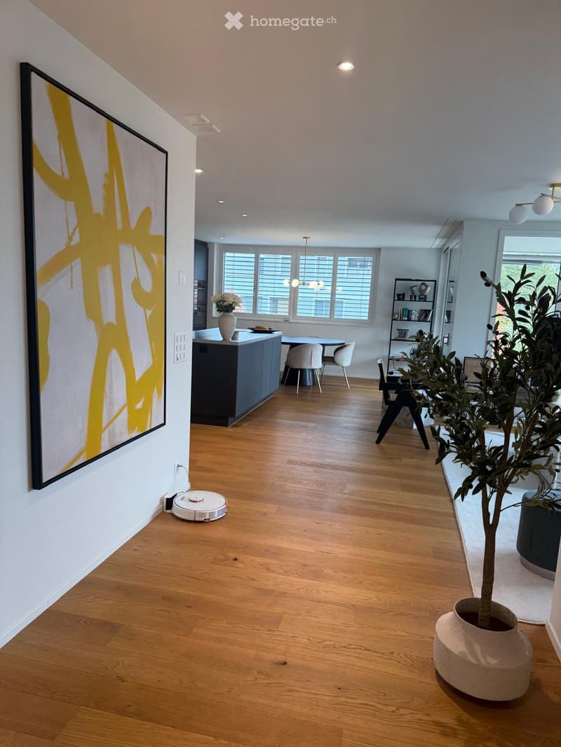 Open-plan living and dining area with wooden flooring, large windows, modern furniture, and decorative elements including a large abstract painting and a potted plant.