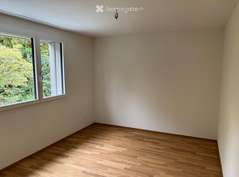 Empty room with wooden flooring, white walls, and large windows offering a view of greenery outside.